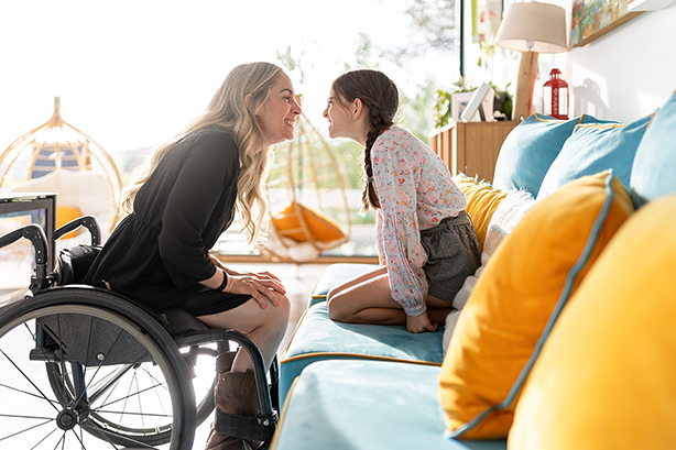 Woman in wheelchair sitting nose-to-nose with a seated young girl, both smiling.
